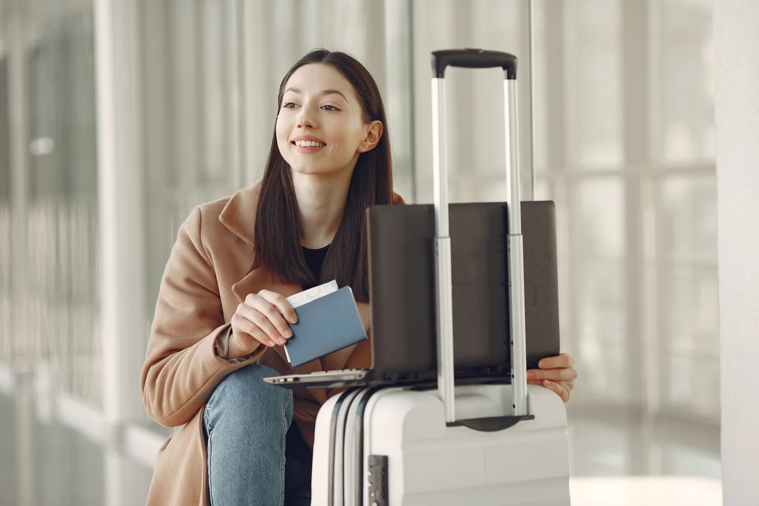 A traveller holding a passport and using a laptop beside luggage